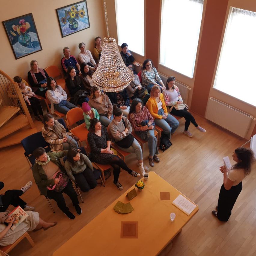 Ein Raum mit vielen Menschen, die auf Stühlen sitzen. Eine Frau steht vor einem Tisch mit Papieren darauf. Eine Kronleuchter hängt von der Decke.