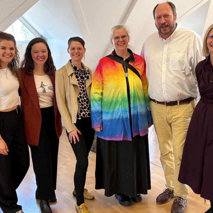 Eine Gruppe von sechs Menschen, drei Frauen und drei Männer, lächeln und stehen zusammen in einem Raum. Sie tragen formelle Kleidung.