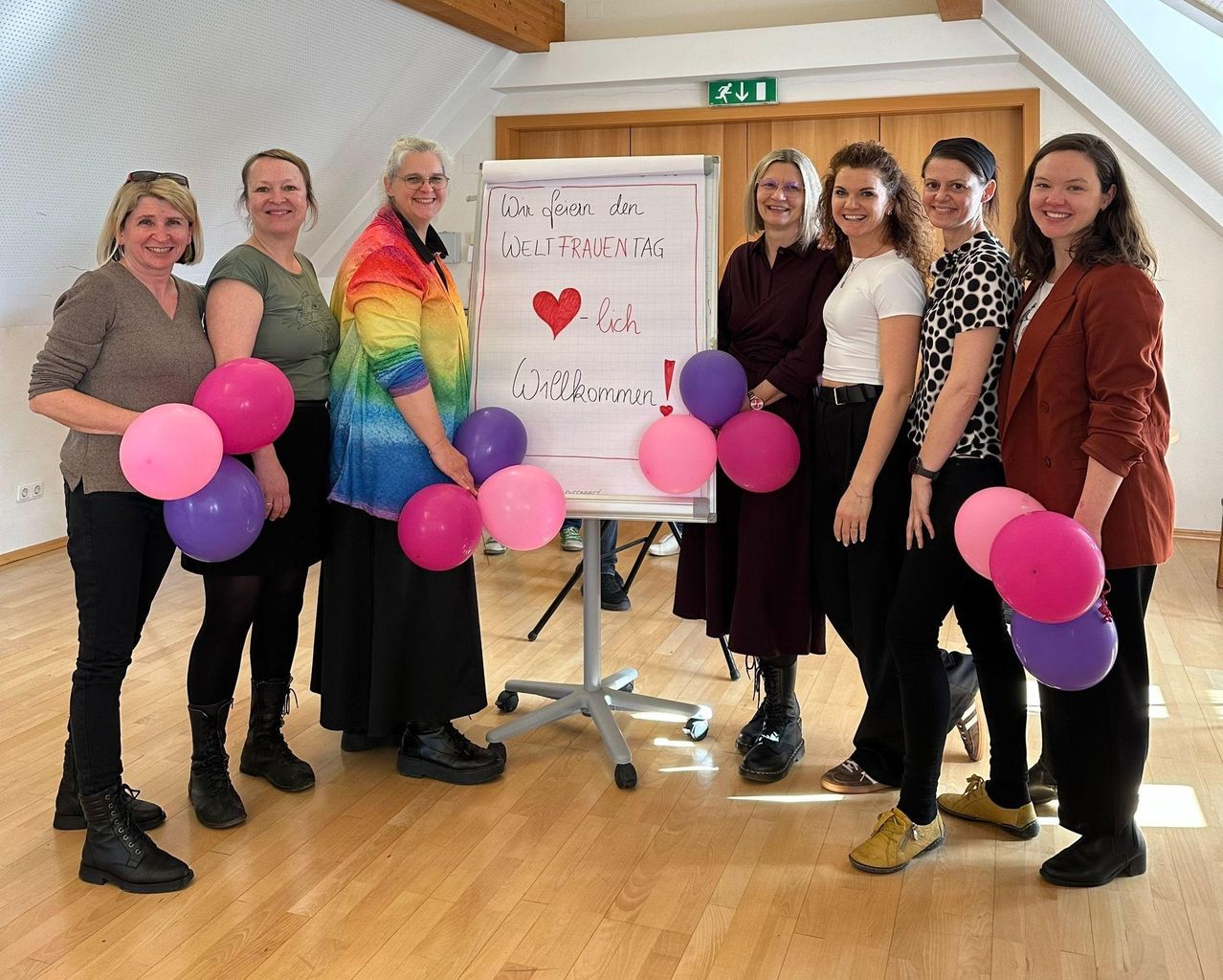 Eine Gruppe von Frauen steht in einem Raum und hält Luftballons. Ein Whiteboard hinter ihnen hat 'Wir freuen den Weltfrauentag' und 'Willkommen!' in Rot geschrieben. Sie alle lächeln.