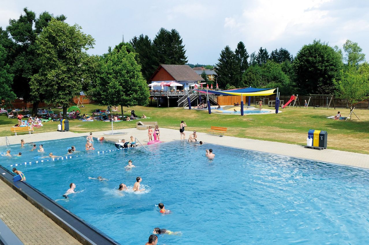 Ein großer Außenpool mit Schwimmern und Leuten, die sich auf der Wiese entspannen. Im Hintergrund befinden sich Bäume, ein Gebäude und eine Rutsche.