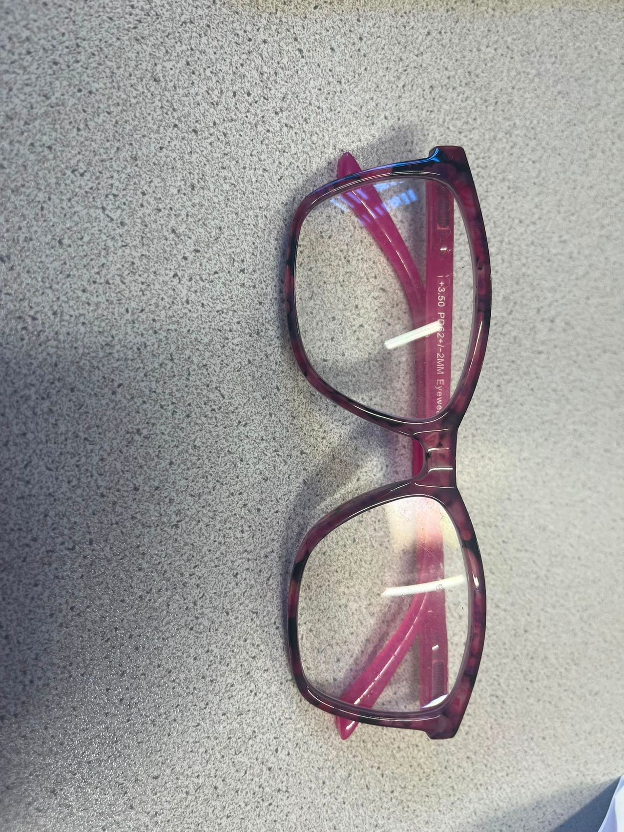 A pair of purple-framed glasses with clear lenses, resting on a textured gray surface. The lenses have a slight gradient tint.