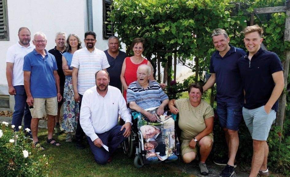 Eine Gruppe von Menschen posiert für ein Foto im Garten, wobei ein älterer Mann im Rollstuhl sitzt.