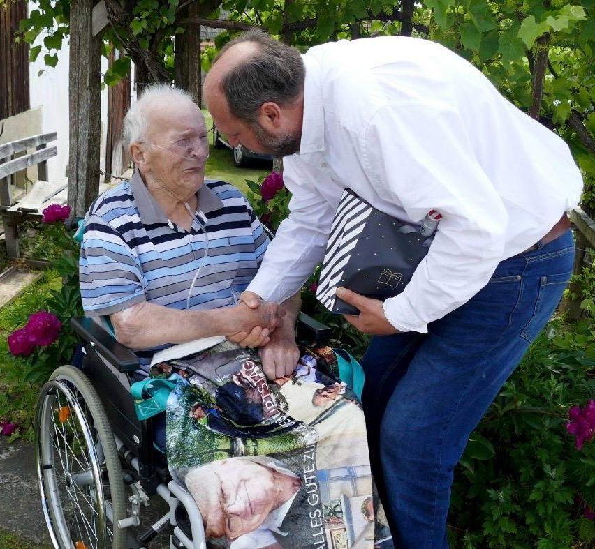 Ein älterer Mann im Rollstuhl hält die Hand eines Mannes in einem weißen Hemd. Der ältere Mann hat eine Fotodecke mit einem Bild von ihm darauf.