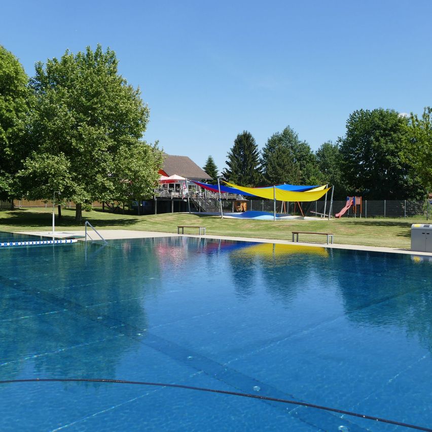 Ein großer Swimmingpool, umgeben von einer Rasenfläche mit bunten Zelten und einem Spielplatz in der Ferne unter einem klaren Himmel.