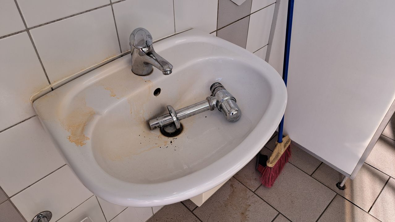 A dirty sink with a silver faucet and a drain pipe inside, next to a broom on tiled floor.