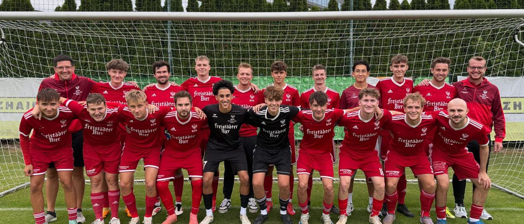 Eine Fußballmannschaft posiert vor einem Tor für ein Foto, mit roten Trikots, auf denen 'Freistadler Bier' steht. Ein Gebäude und Bäume sind im Hintergrund zu sehen.