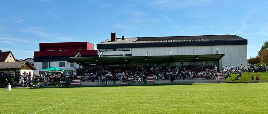 Eine große Menschenmenge versammelt sich auf den Tribünen eines Stadions, um eine Sportveranstaltung bei blauem Himmel mit einzelnen Wolken zu verfolgen. Das Spielfeld ist gut gepflegt und das Stadion hat ein modernes Design.