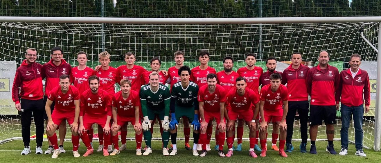 Eine Fußballmannschaft in roten Trikots posiert für ein Foto vor einem Tor, wobei ein Spieler in Grün gekleidet ist.