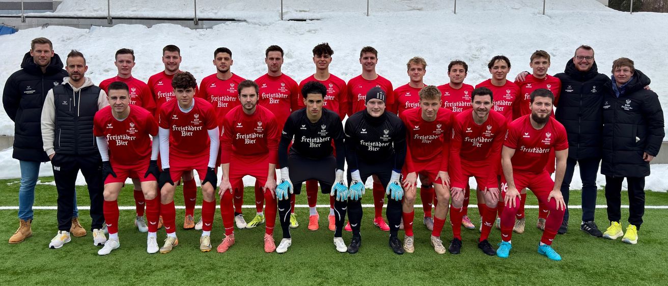 Eine Fußballmannschaft in roten Trikots und Handschuhen posiert für ein Foto auf einem verschneiten Feld mit einem Gebäude im Hintergrund.