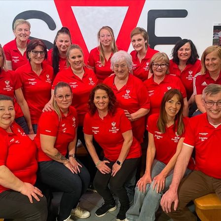 Eine Gruppe von Frauen und Männern in roten Shirts lächelt und posiert für ein Foto. Sie stehen vor einer weißen Wand mit den Buchstaben SVE. Sie sind wahrscheinlich ein Team.