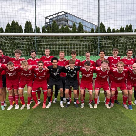 Eine Fußballmannschaft in roten Trikots posiert für ein Foto vor einem Tor. Sie lächeln und scheinen zu feiern.