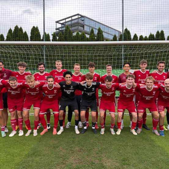 Eine Fußballmannschaft in roten Trikots posiert für ein Foto vor einem Tor. Sie lächeln und scheinen zu feiern.