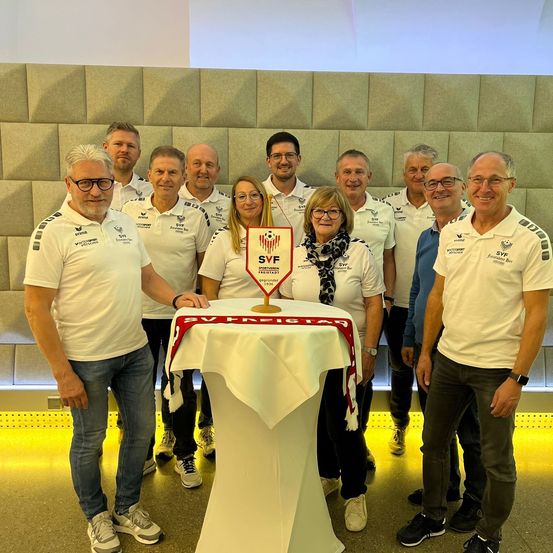 Eine Gruppe von Menschen in weißen Hemden und mit Brillen posiert für ein Foto vor einer Wand. Sie lächeln und scheinen zu feiern. Eine Frau steht in der Mitte, und ein Tisch mit weißer Tischdecke steht vor ihnen.