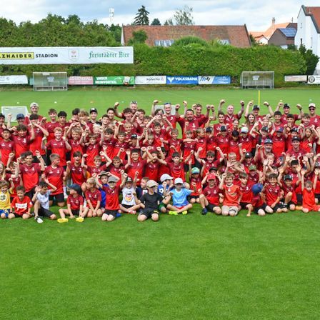 Eine Gruppe junger Fußballspieler, die rote Trikots tragen, posiert für ein Teamfoto auf einem grünen Feld mit einem Banner im Hintergrund.