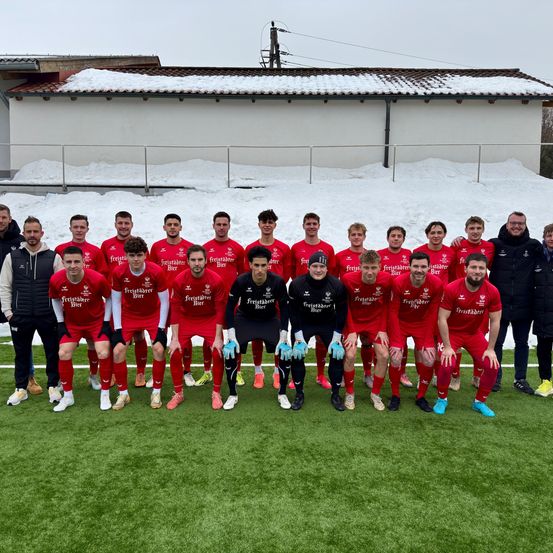 Eine Fußballmannschaft in roten Trikots posiert für ein Foto auf einem verschneiten Feld, zwei Trainer in Schwarz. Dahinter ein Gebäude mit Schnee auf dem Dach.