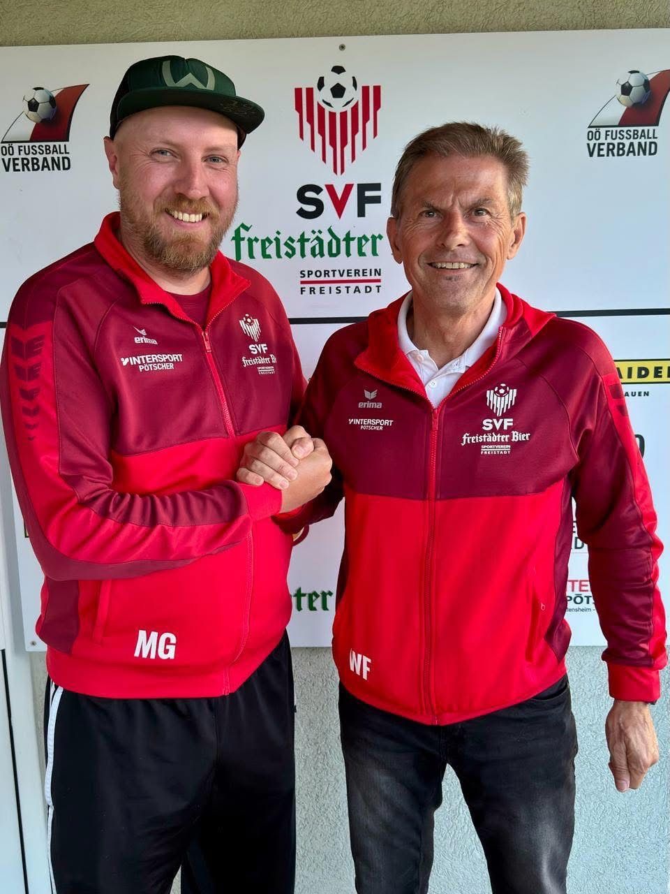 Two men in red jackets with Intersport logos, standing in front of a wall with sports team branding. One wears a black cap, the other a white shirt. They appear to be smiling and posing for a photo.