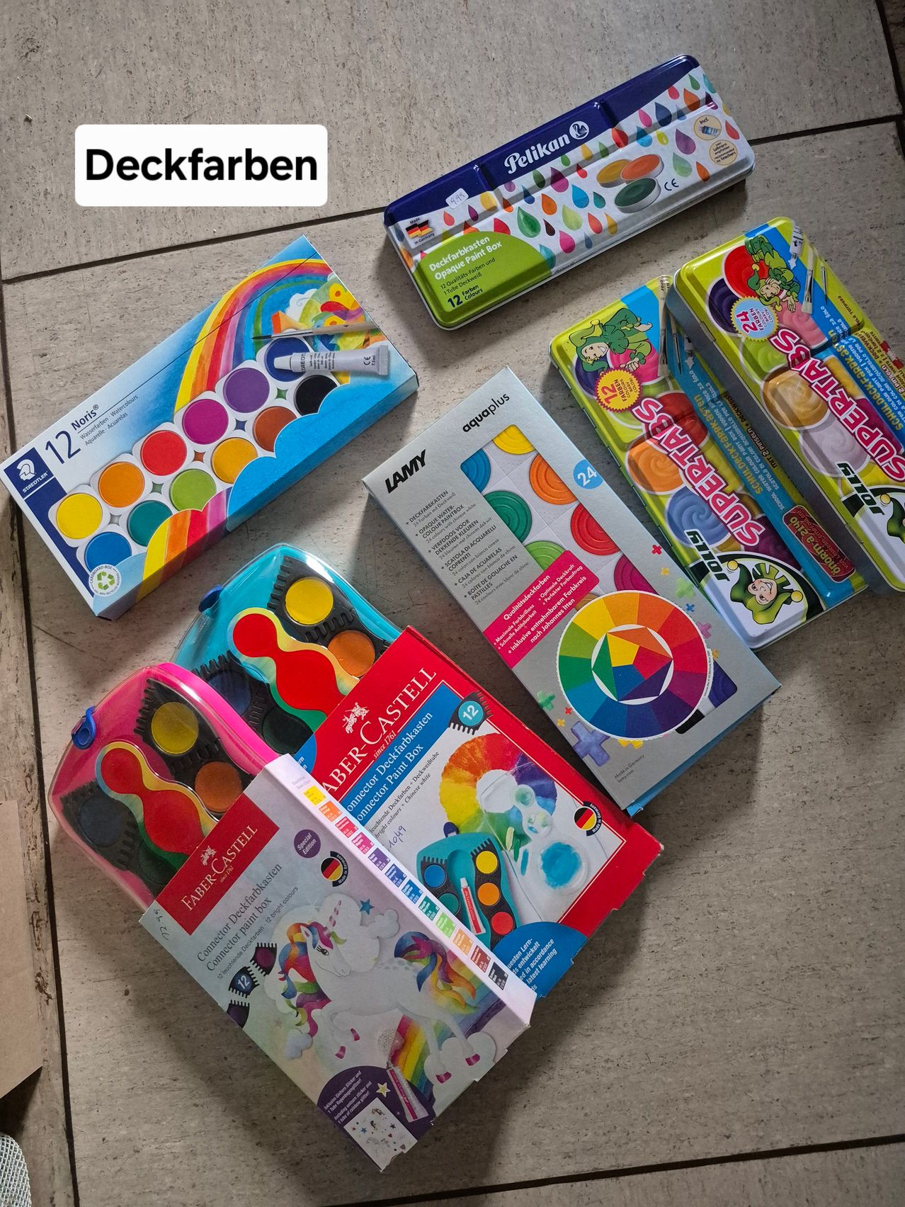 Verschiedene Schachteln mit Deckfarben und Malsets sind ausgestellt. Dazu gehören eine Schachtel mit 12 Aquarellen, eine Schachtel mit Regenbogendesign und eine Schachtel mit Einhorndesign.