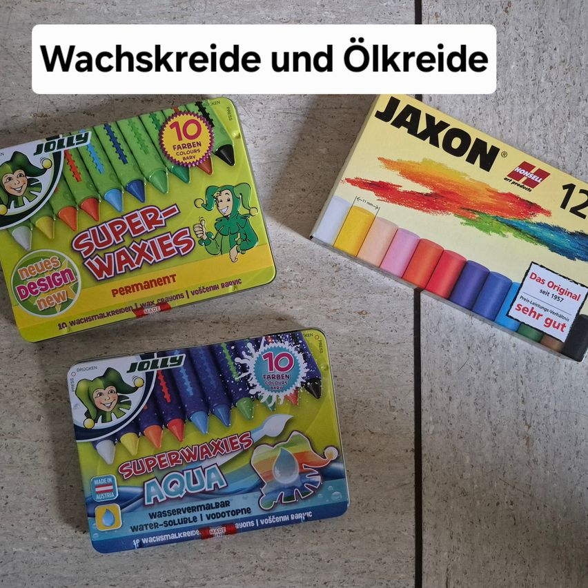 Drei Schachteln mit Wachsmalstiften auf einer Oberfläche. Die linke Schachtel hat einen Joker darauf und steht 'Super-Waxies Permanent'. Die rechte Schachtel hat 'Jaxon' darauf geschrieben. Die mittlere Schachtel sagt 'SuperWaxies Aqua'.