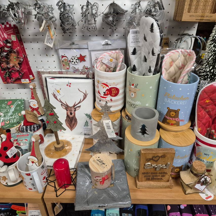 Ein Holzregal zeigt verschiedene Weihnachtsartikel, darunter Tassen, Holzkisten und Dekorationen. Einige Artikel haben Weihnachtsmann-Designs, während andere Rehe, Weihnachtsbäume und Herzen zeigen. An der Wand hängen auch einige Backwerkzeuge.