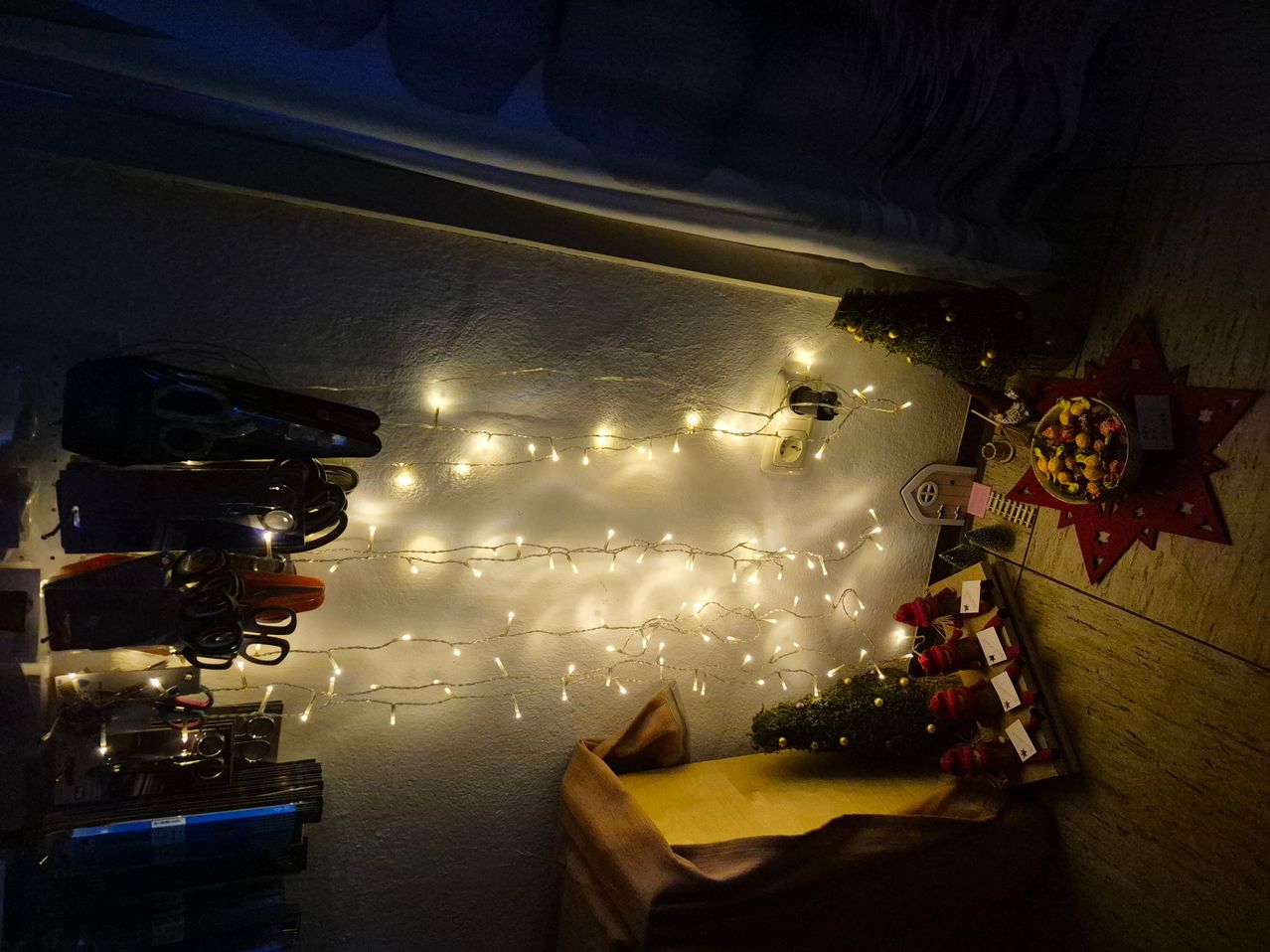 Ein Zimmer mit weißen Wänden, einer beleuchteten Lichterkette, einem kleinen Weihnachtsbaum und einer Obstschüssel auf einem Tisch.