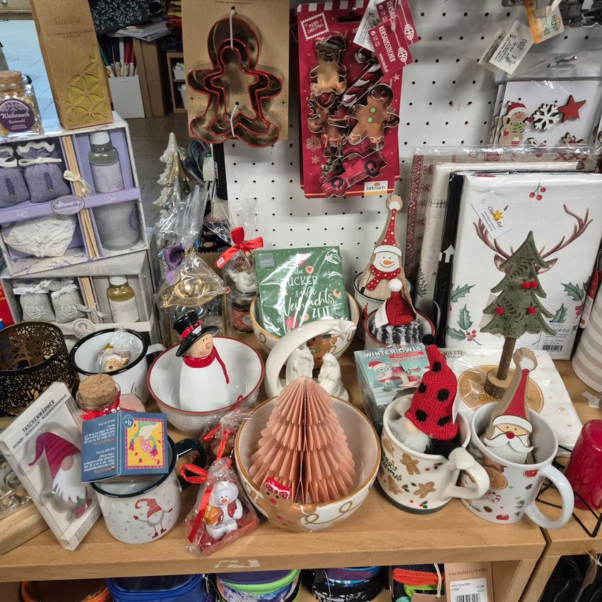 Ein hölzerne Regalplatte zeigt eine Vielzahl von Feiertagsartikeln, darunter Tassen, Ornamente und Geschenkpapier. Bemerkenswert sind ein Korb mit einem Weihnachtsbaum und eine Schachtel mit Weihnachtskarten.