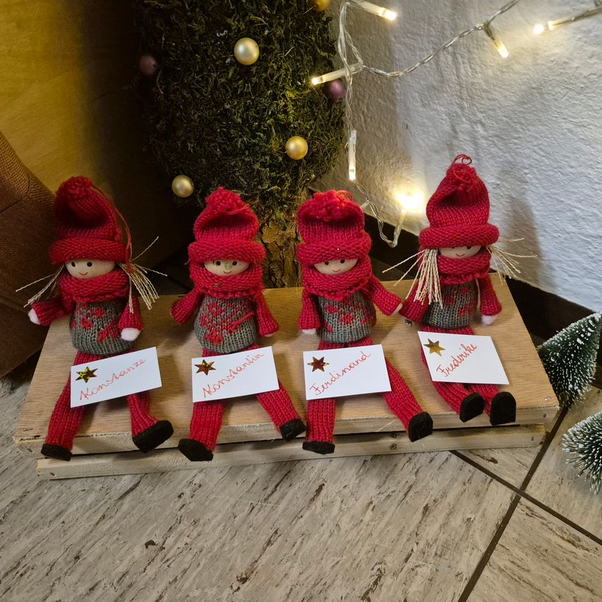 Vier gestrickte Weihnachtselfen mit roten Mützen und Schals stehen auf einem Holzbrett. Jede Figur hat ein Namensschild. Ein geschmückter Weihnachtsbaum und Lichter sind im Hintergrund.