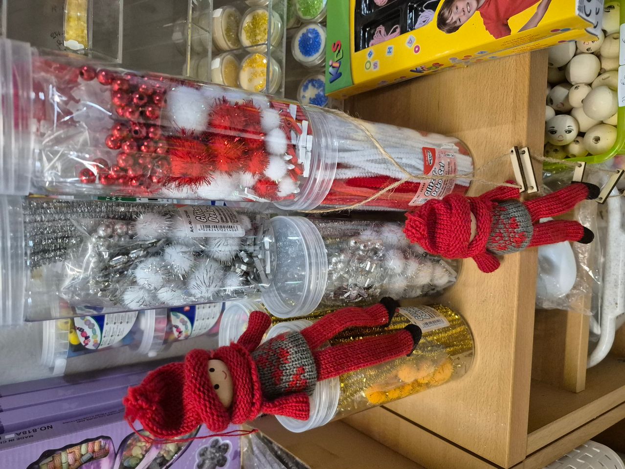 Eine Ausstellung von Bastelmaterialien auf einem Holzregal, einschließlich Röhren mit roten und weißen Perlen, roten und grauen gestrickten Puppen und einer gelben Schachtel mit einem Bild eines Kindes.