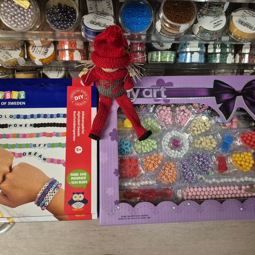 Eine Ausstellung mit zwei farbenfrohen DIY-Bastelsets: eines mit Alphabetperlen und ein weiteres mit verschiedenen Perlen in unterschiedlichen Farben. Eine Puppe mit rotem Hut sitzt auf dem Perlenset.