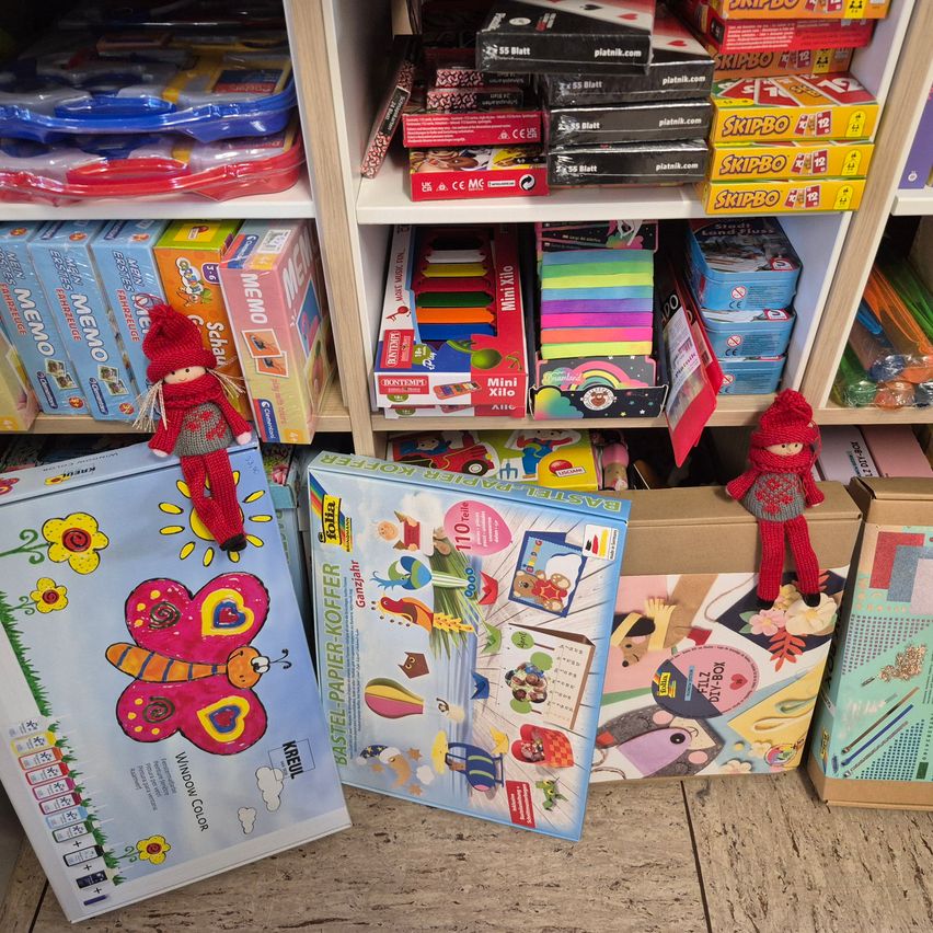 Ein Spielzeuggeschäft mit einem Holzboden und Regalen, die mit verschiedenen Spielzeugen gefüllt sind, darunter Bastelarbeiten, Brettspiele und Puzzles. Zwei geöffnete Kisten, eine mit Schmetterlingsdesign und die andere mit einem bunten Puzzle.