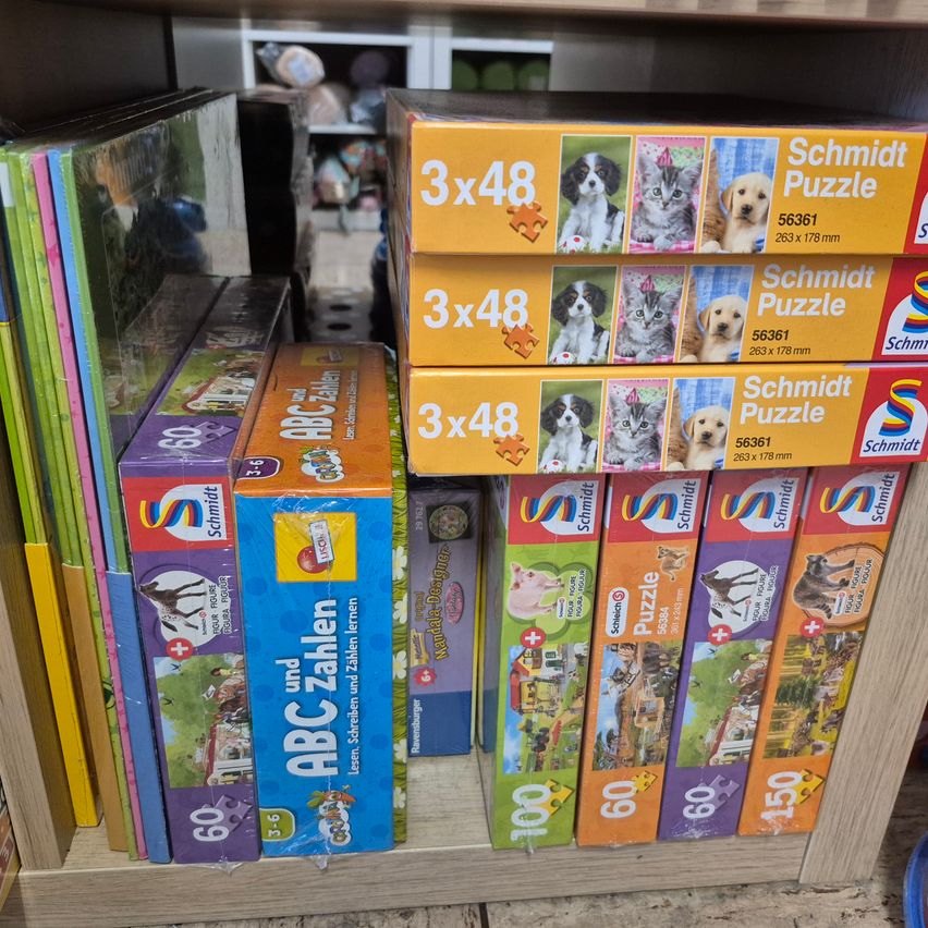 Ein Regal zeigt verschiedene Puzzleboxen; einige sind Schmidt Puzzles, andere haben ABC Zahlen. Einige Puzzles zeigen Tiere wie Hunde und Katzen, während andere Landschaften und Bauernhoftiere darstellen.