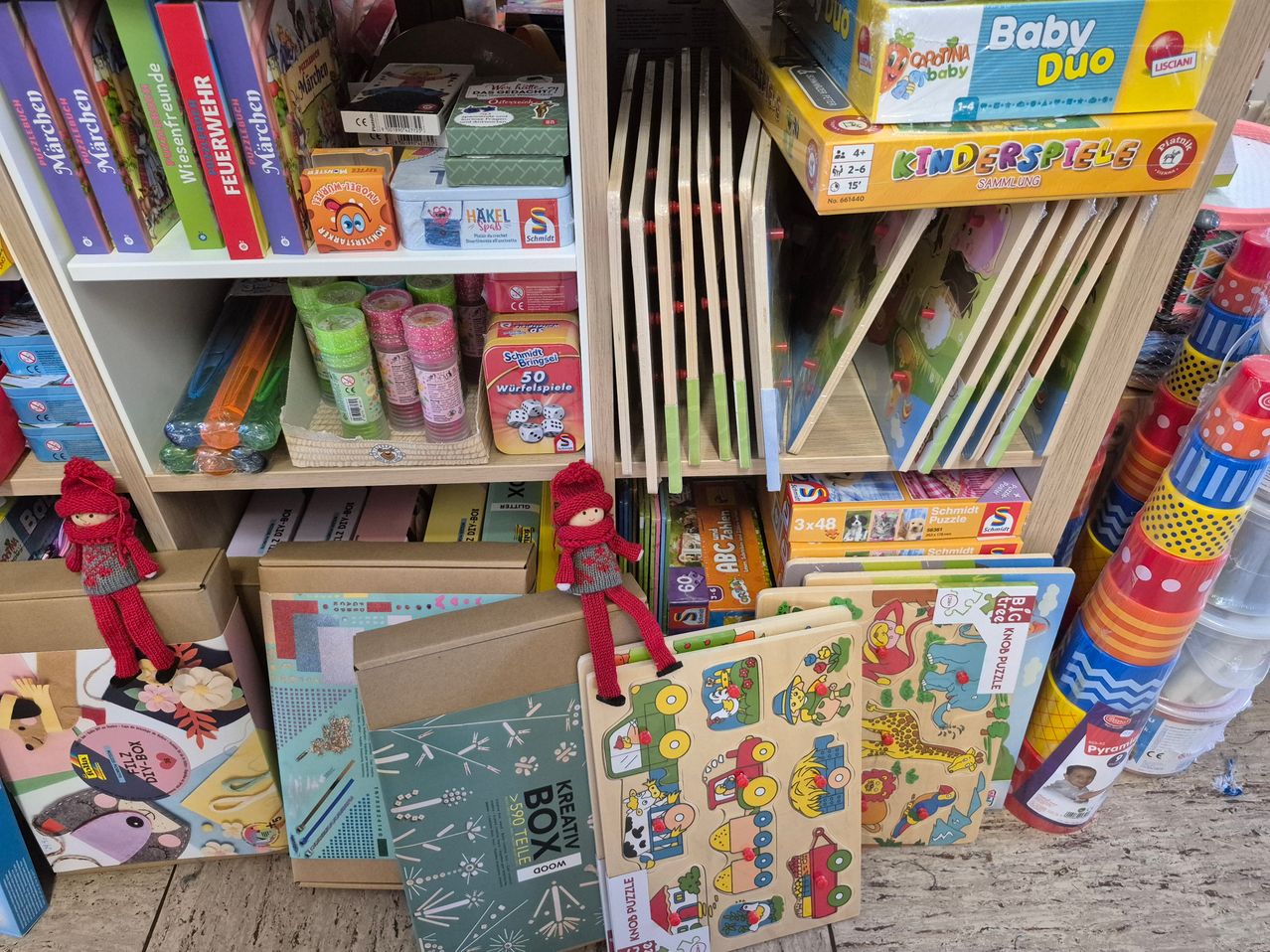 Ein Spielzeuggeschäft gefüllt mit verschiedenen Spielen und Spielzeugen. Eine Puppe sitzt auf einem Beutel im Vordergrund. Regale sind mit bunten Schachteln, Puzzles und Malutensilien gestapelt.