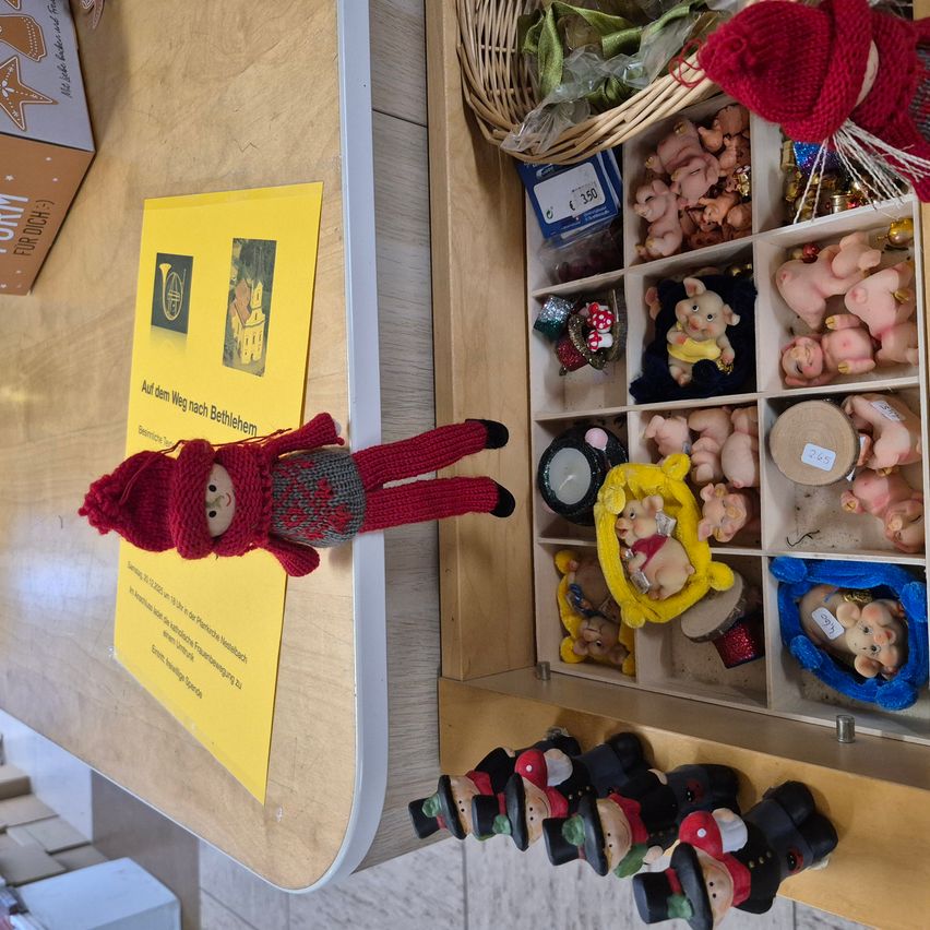 Ein Holztisch zeigt eine Sammlung von Spielzeug, darunter eine gestrickte rote Puppe, einen Korb mit verschiedenen Gegenständen und ein Holztablett mit Schweinespielzeug. Ein gelbes Papier mit Text ist auf der linken Seite angeheftet.