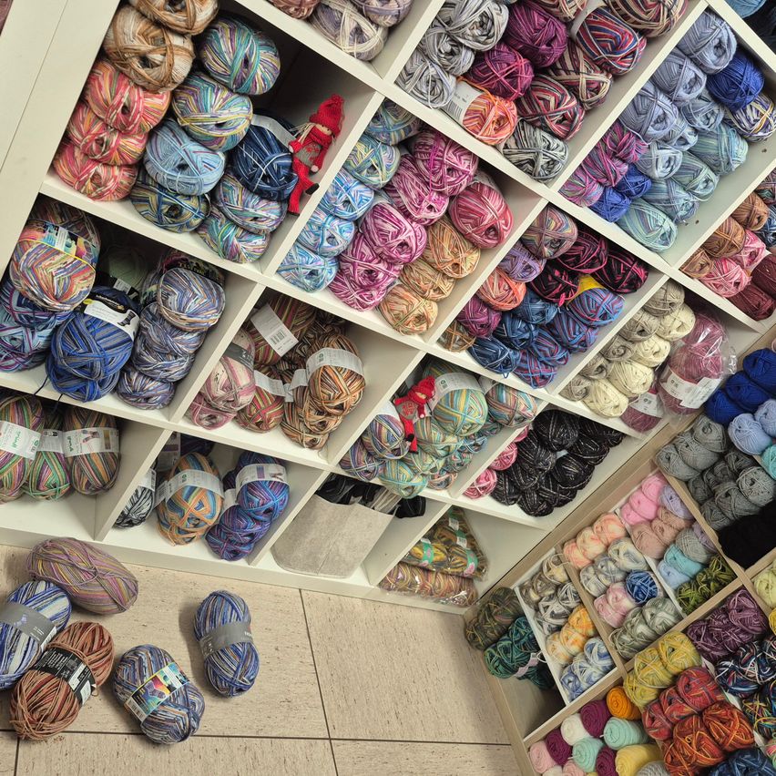Ein Garnladen zeigt mehrere Regale mit Garnkugeln in verschiedenen Farben. Einige Garnkugeln liegen auf dem Boden, und eine Puppe ist auf einem Regal.