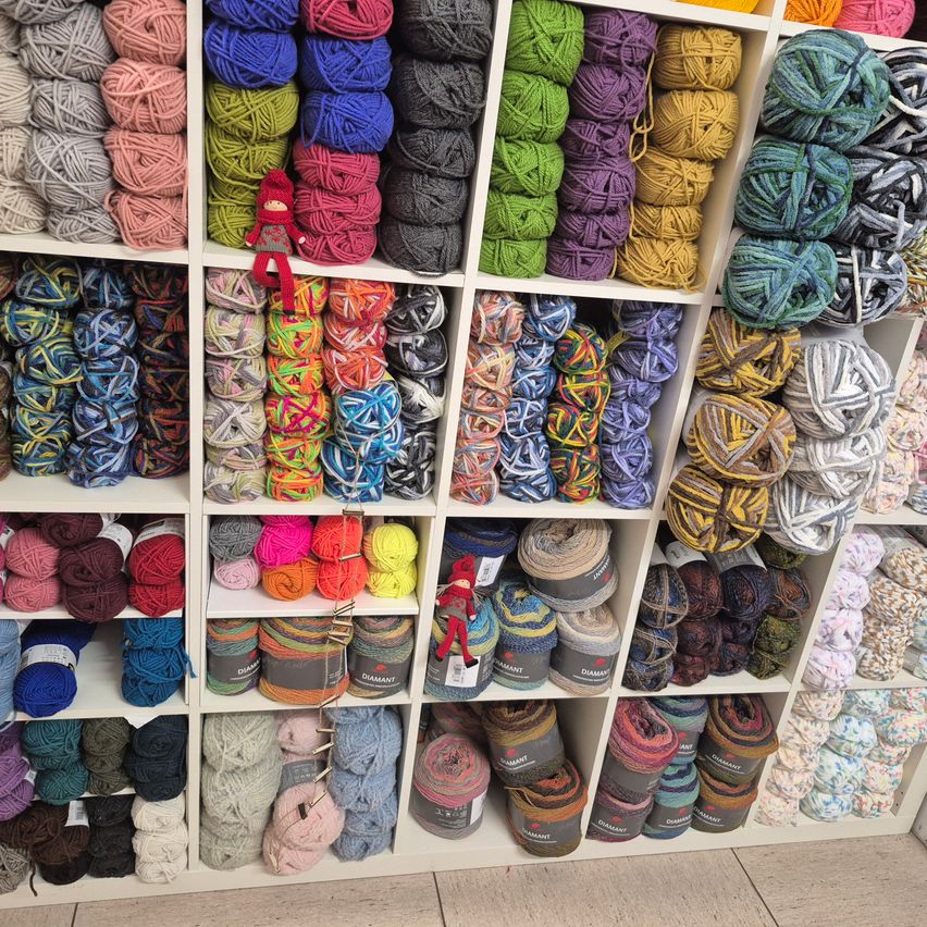 Eine Ausstellung von farbenfrohen Garnknäueln auf Regalen, einschließlich Rosa-, Blau-, Grün-, Lila- und weiteren Farbtönen. Eine kleine Figur sitzt auf einem Stapel Garn.