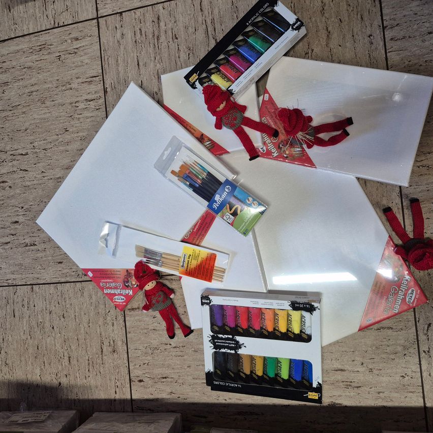 Eine Auswahl an Malutensilien auf einer Holzoberfläche. Es enthält zwei rote Elfenfiguren, zwei Sets Malpinsel, zwei Sets Aquarelle, ein Set Buntstifte und mehrere leere Leinwände. Die Szene wird von einem hellen Licht beleuchtet.