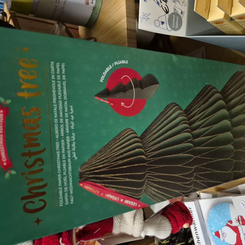 Eine grüne Box mit dem Text 'Weihnachtsbaum'. Ein gefalteter Papierweihnachtsbaum ist abgebildet, mit einem roten Kreis auf der Vorderseite. Darunter befinden sich einige Weihnachtsdekorationen.