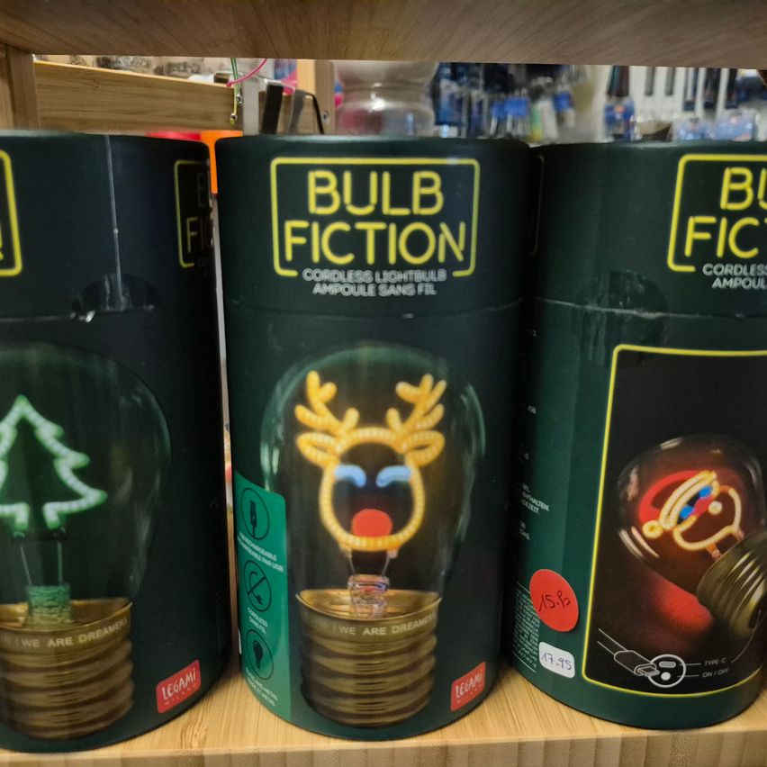 Drei grüne zylindrische Boxen mit den Worten Bulb Fiction sind ausgestellt. Die mittlere Box zeigt eine leuchtende Glühbirne mit einem leuchtenden Rentier darauf. Die Boxen stehen auf einem Holzregal.