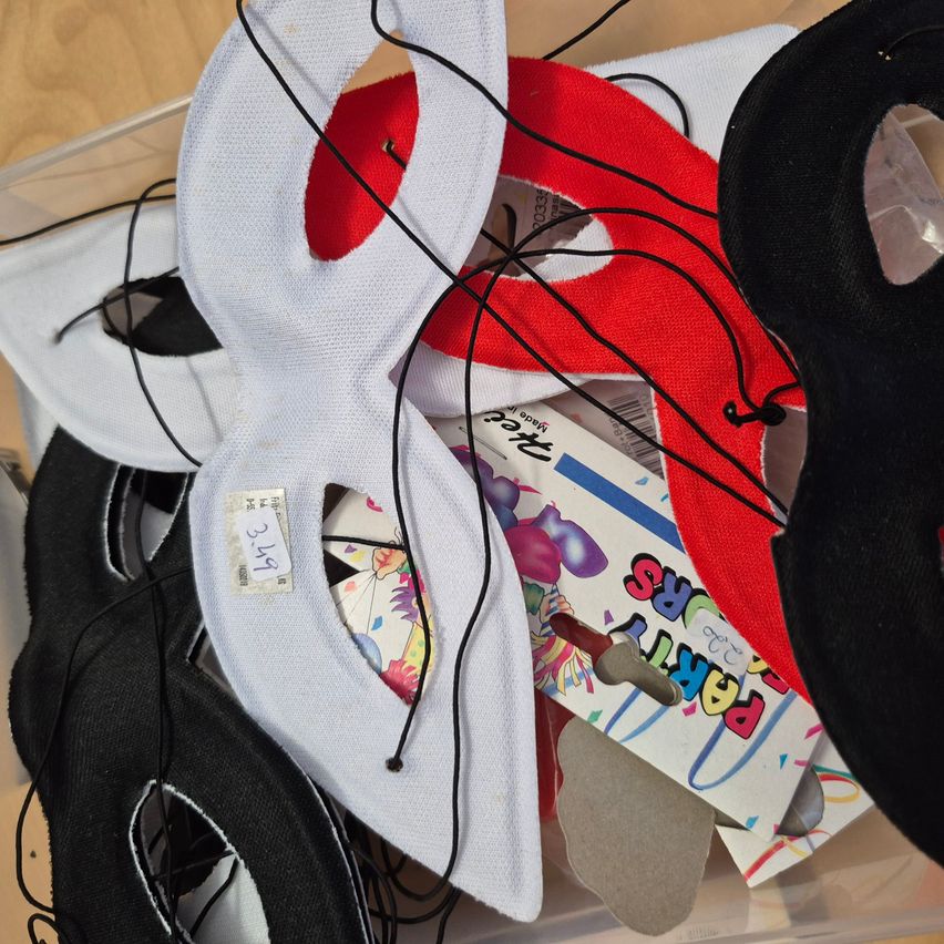 Verschiedene Partymasken in Schwarz, Weiß und Rot sind in einem durchsichtigen Plastikbehälter ausgestellt. Ein farbenfrohes Karton mit dem Wort 'Party' ist ebenfalls sichtbar.
