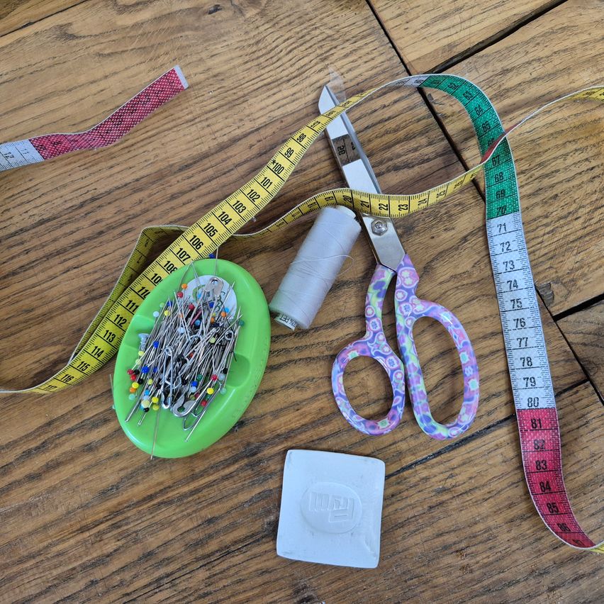 Eine Sammlung von Nähwerkzeugen auf einer Holzoberfläche: ein Maßband, Schere, ein Fadenspule, ein Nadelkissen und ein Stück Stoff.