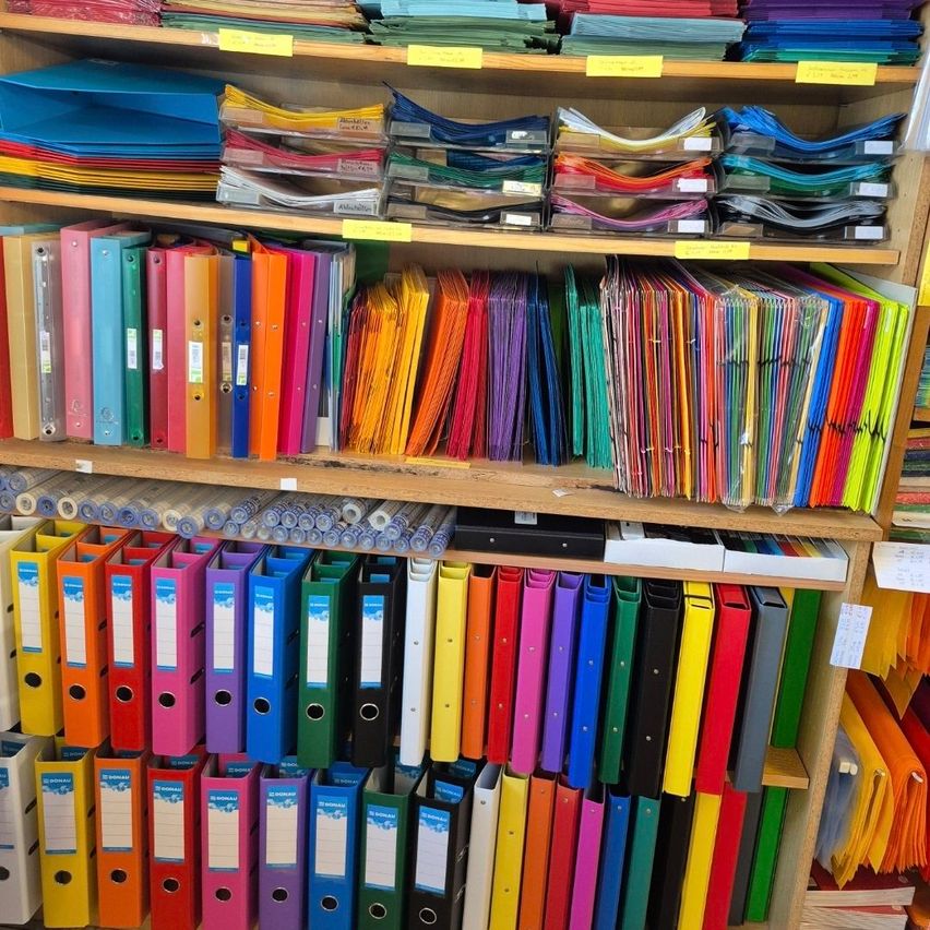 Ein Holzregal gefüllt mit verschiedenen farbigen Ordnern und Mappen. Einige Ordner haben Etiketten, während andere leer sind. Über den Ordnern befinden sich Stapel farbiger Papiere. Das Regal ist ordentlich organisiert.
