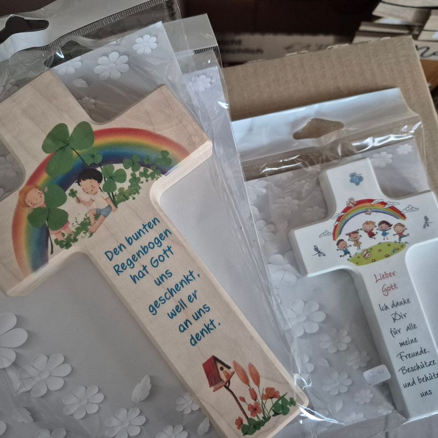 Zwei Holzkreuze in Plastikverpackung. Eines zeigt einen Regenbogen, Kinder und ein Haus mit einem Zitat. Das andere hat ein ähnliches Design mit anderer Beschriftung.