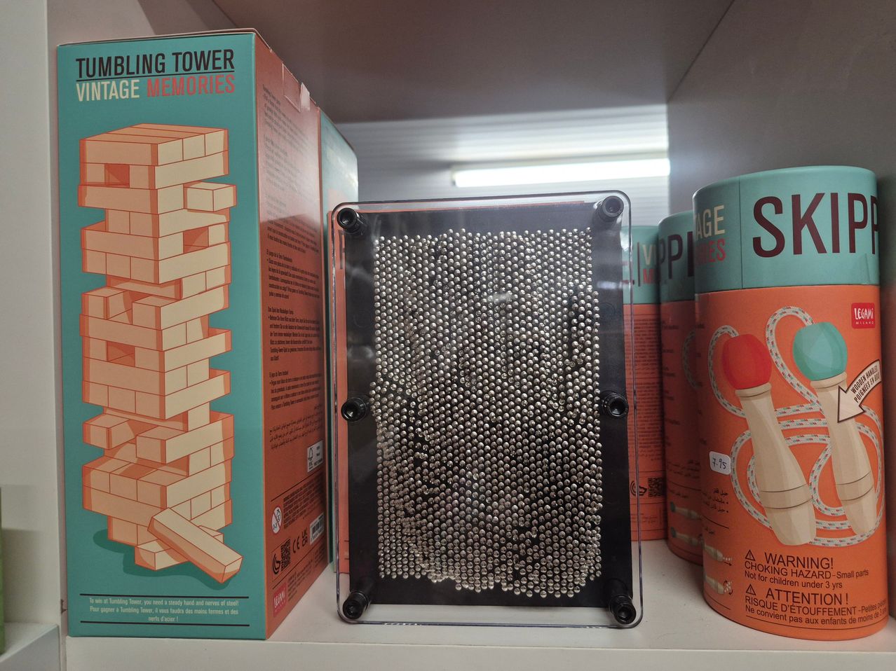 Ein Regal zeigt ein durchsichtiges Gehäuse mit einem Magnetbrett und metallischen Kugeln, neben einer Schachtel für ein Stapelturmspiel und einem Behälter mit rotem Griff.