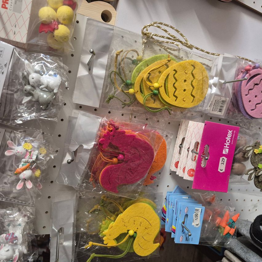 Eine Sammlung von Osterdekorationen und Spielzeug auf einem weißen Pegboard. Die Artikel umfassen Filzeier, Kaninchen, Karotten und andere kleine Spielzeuge, alles in Plastik verpackt. Jeder Artikel hängt an einem Haken, und es gibt Preisschilder.