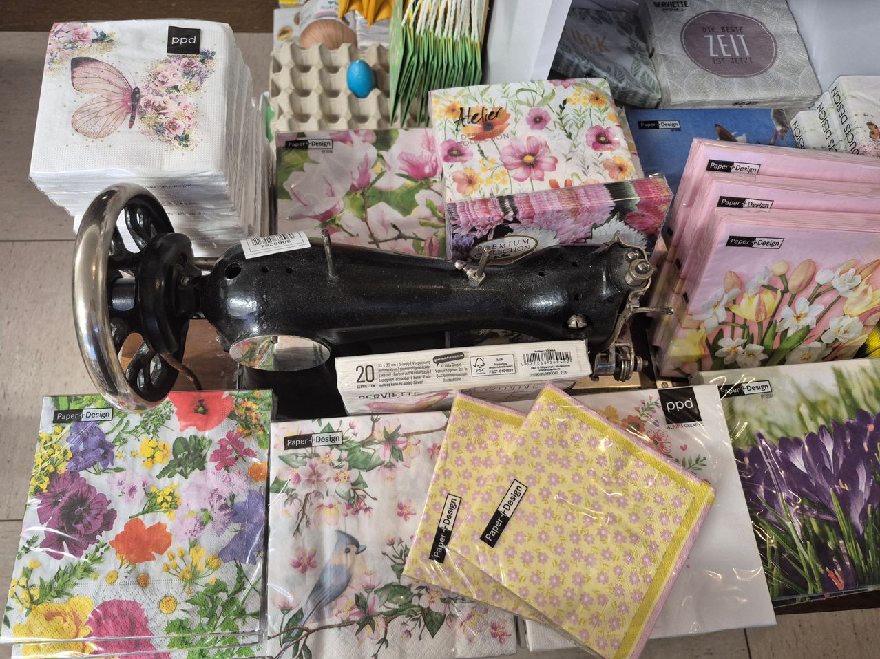 Auf einem Tisch sind verschiedene Gegenstände zu sehen, darunter eine Nähmaschine, Stapel von Servietten mit Blumendesigns, eine Schachtel Papierhandtücher und ein Osterei.