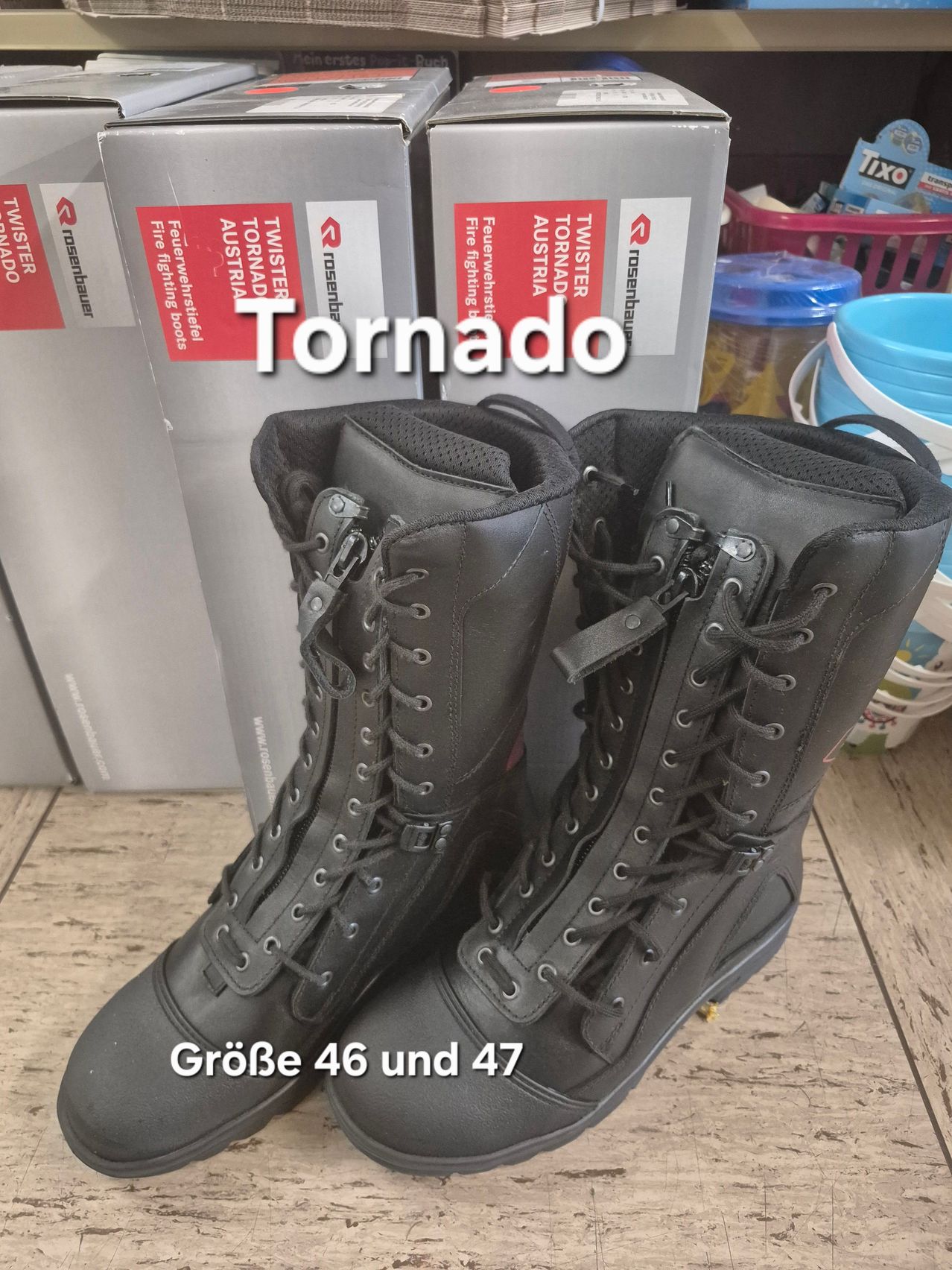 Schwarze Leder-Feuerwehrstiefel mit der Aufschrift 'Tornado' und den Größen 46 und 47. Die Stiefel haben mehrere Reißverschlüsse und Schnürsenkel und stehen auf einer Holzfläche. Dahinter befinden sich Kartons mit 'TWISTER', 'Austria' und 'Feuerwehrstiefel'.
