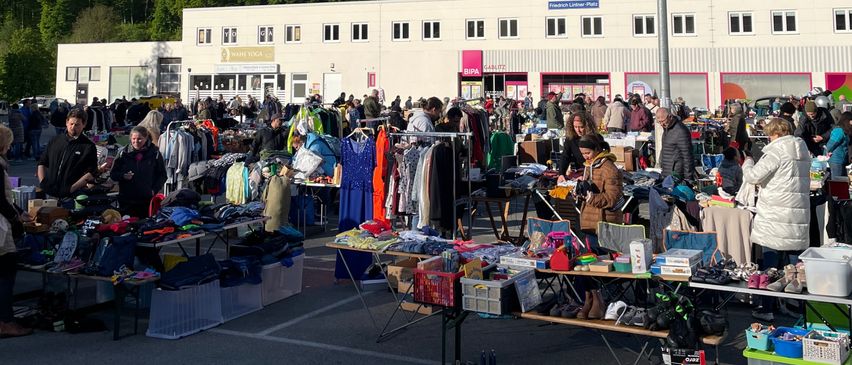 Ein Outdoor-Flohmarkt mit zahlreichen Ständen und Tischen unter einem bewölkten Himmel, mit einem großen Gebäude und einem Hügel im Hintergrund.