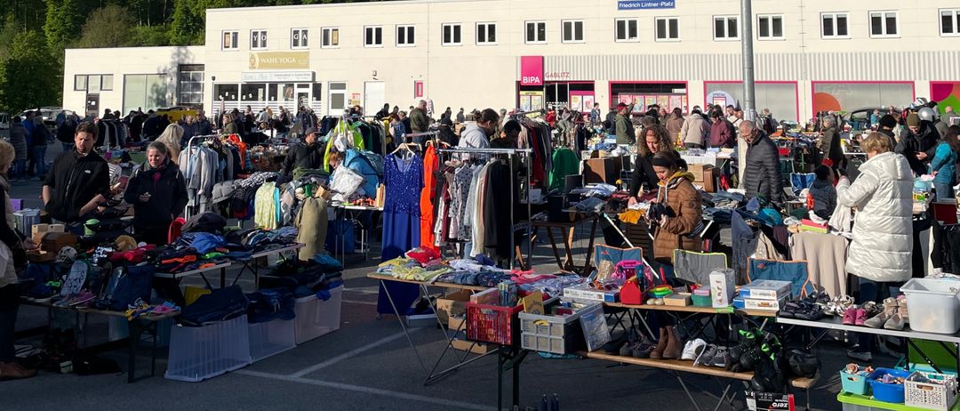 Ein Outdoor-Flohmarkt mit zahlreichen Ständen und Tischen unter einem bewölkten Himmel, mit einem großen Gebäude und einem Hügel im Hintergrund.