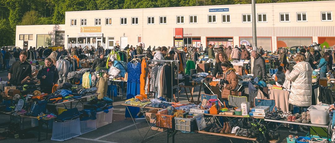 Ein geschäftiger Flohmarkt mit zahlreichen Ständen, die verschiedene Artikel unter einem bewölkten Himmel verkaufen. Im Hintergrund ist ein großes Gebäude mit mehreren Fenstern zu sehen.
