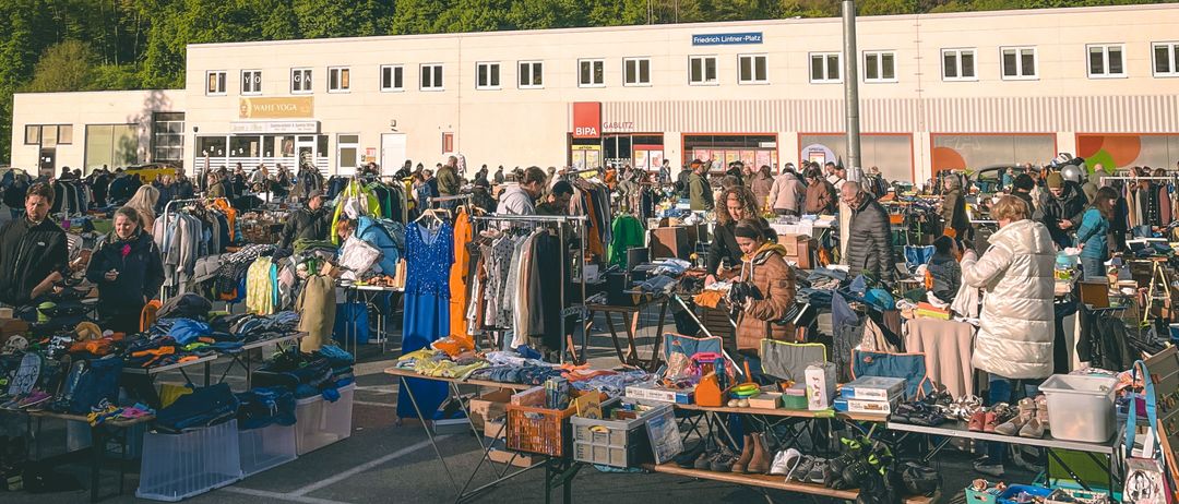 Ein großer Flohmarkt ist voller Menschen vor einem Gebäude mit vielen Fenstern, vor dem Hintergrund eines bewölkten Himmels und eines üppig grünen Hügels.