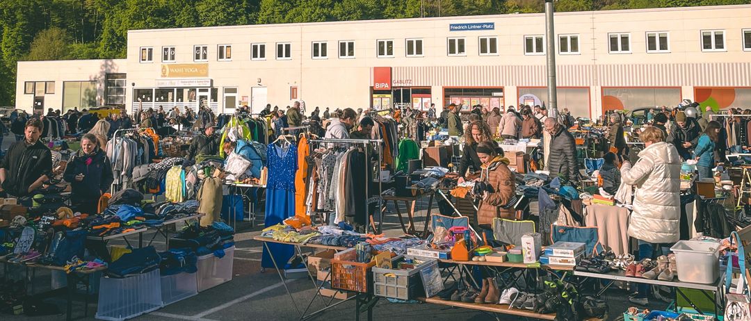 Ein Flohmarkt mit verschiedenen Ständen, Menschen beim Stöbern und Artikeln zum Verkauf. Ein Gebäude mit vielen Fenstern im Hintergrund und eine Straßenlaterne steht in der Mitte.